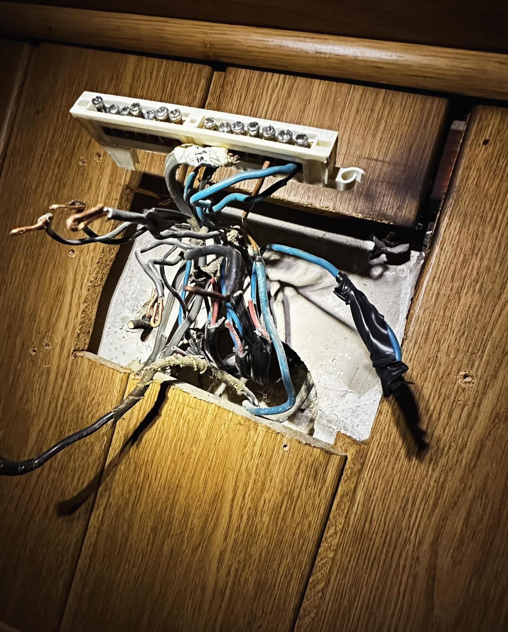 Otwór w boazerii ukazuje splątane przewody elektryczne, częściowo zaizolowane taśmą, podłączone do listwy zaciskowej. Widoczne kolory przewodów: niebieski, czarny, czerwony.