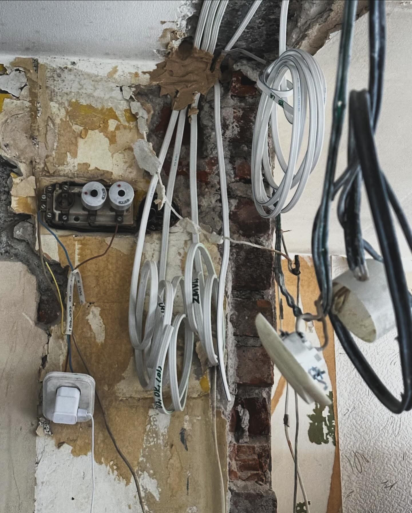 Niefunkcjonująca instalacja elektryczna z przewodami, puszką bezpiecznikową i gniazdkiem na tle zniszczonej ściany w trakcie remontu. Widoczne kable i fragment ceglanej ściany.
