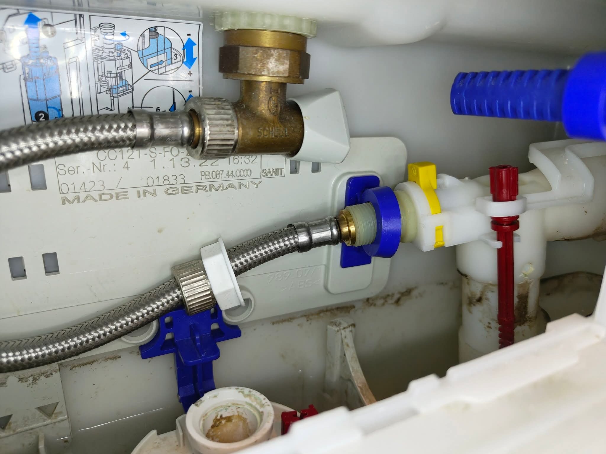 Wnętrze spłuczki WC z widocznymi elementami instalacji wodnej: zawór Schell, wężyk w oplocie stalowym, elementy z tworzywa w kolorach białym, niebieskim i żółtym. Widoczne instrukcje.