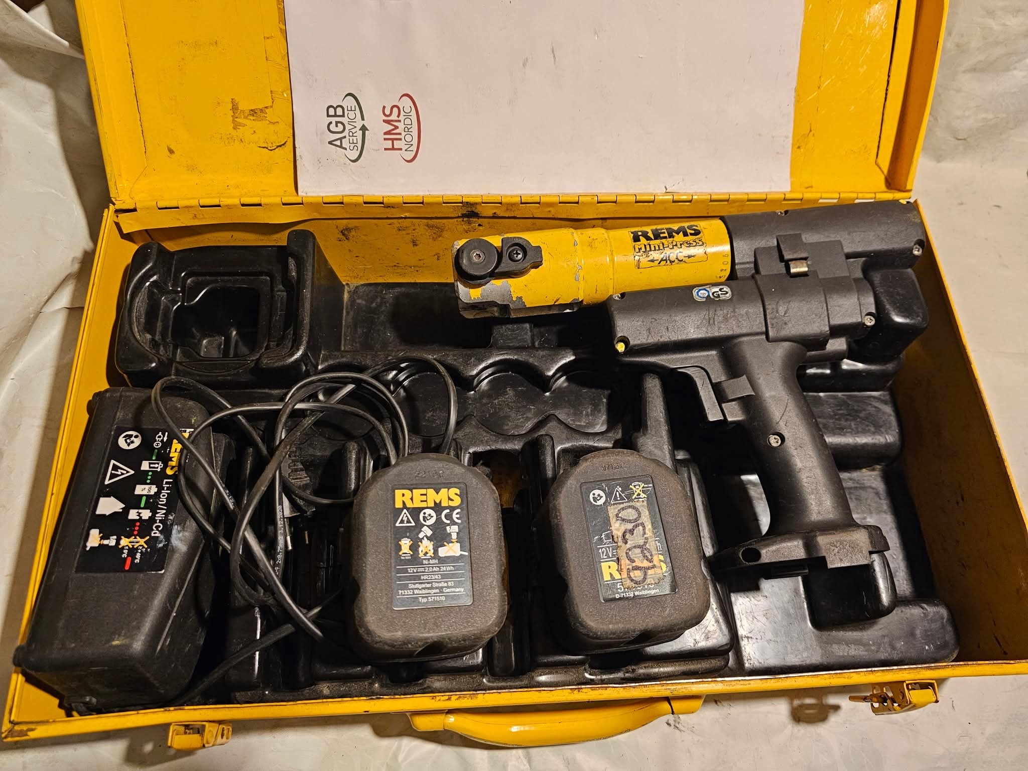 Otwarta żółta walizka narzędziowa z akumulatorową zaciskarką REMS Mini-Press ACC, dwoma akumulatorami i ładowarką. Widoczne logo AGB Service i HMS Nordic.