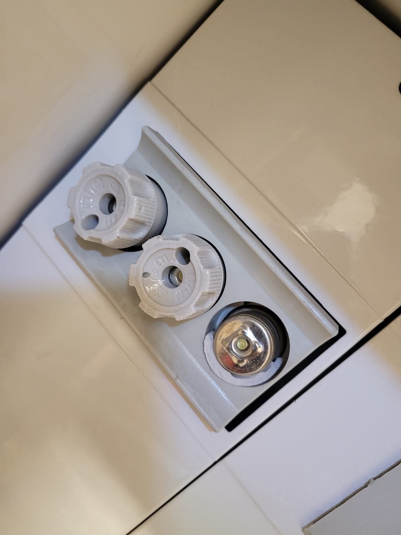 Białe pokrętła termostatyczne grzejnika, dwa z nich założone, a jeden z metalowym trzpieniem widocznym w gnieździe, na tle białej obudowy grzejnika.