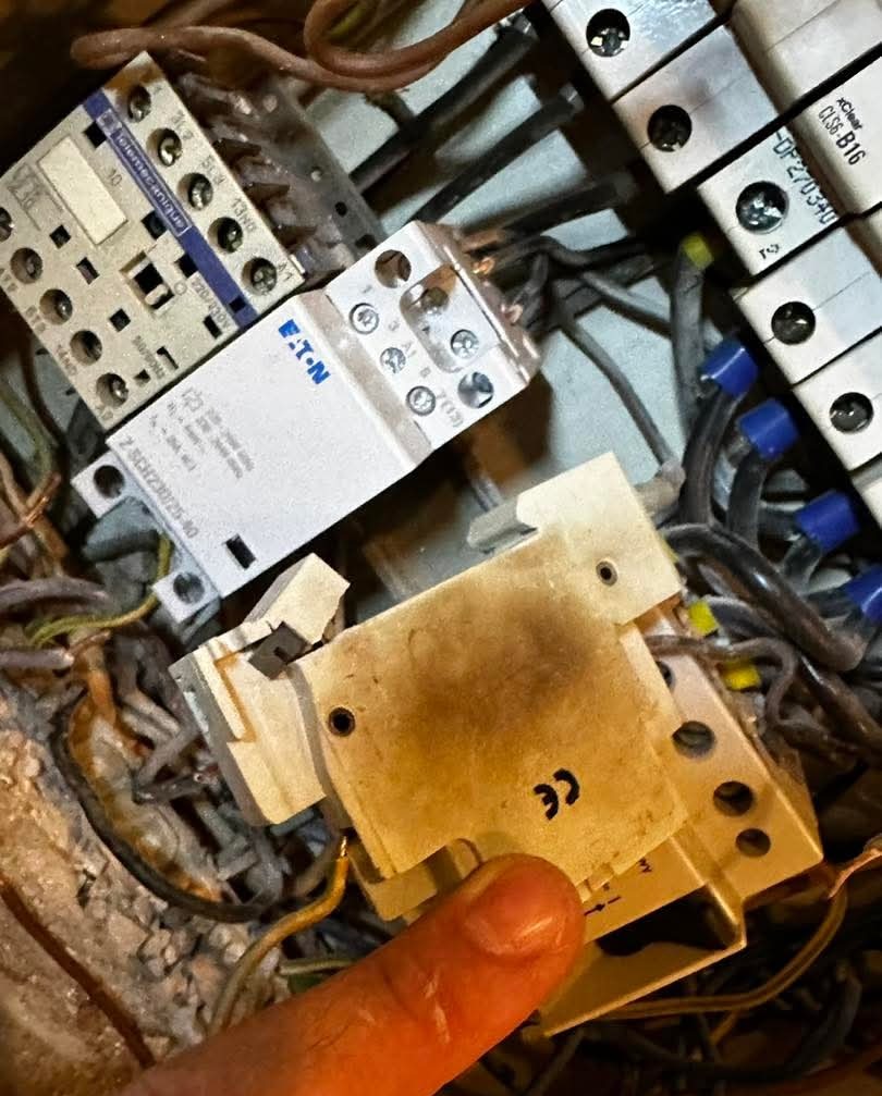 Uszkodzony stycznik EATON z widocznymi śladami przepalenia, w tle inne elementy instalacji elektrycznej i przewody. Palec wskazujący na uszkodzenie.