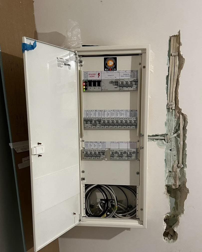 Otwarta skrzynka elektryczna z widocznymi bezpiecznikami i przewodami, zamontowana na ścianie z wyżłobieniem na kable. Widoczne przewody.