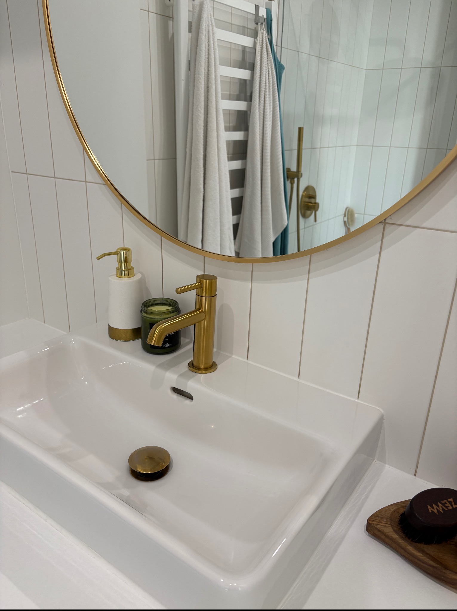 Biała, prostokątna umywalka z złotą baterią i okrągłym lustrem w łazience z białymi płytkami. Widoczny dozownik mydła i świeca zapachowa.