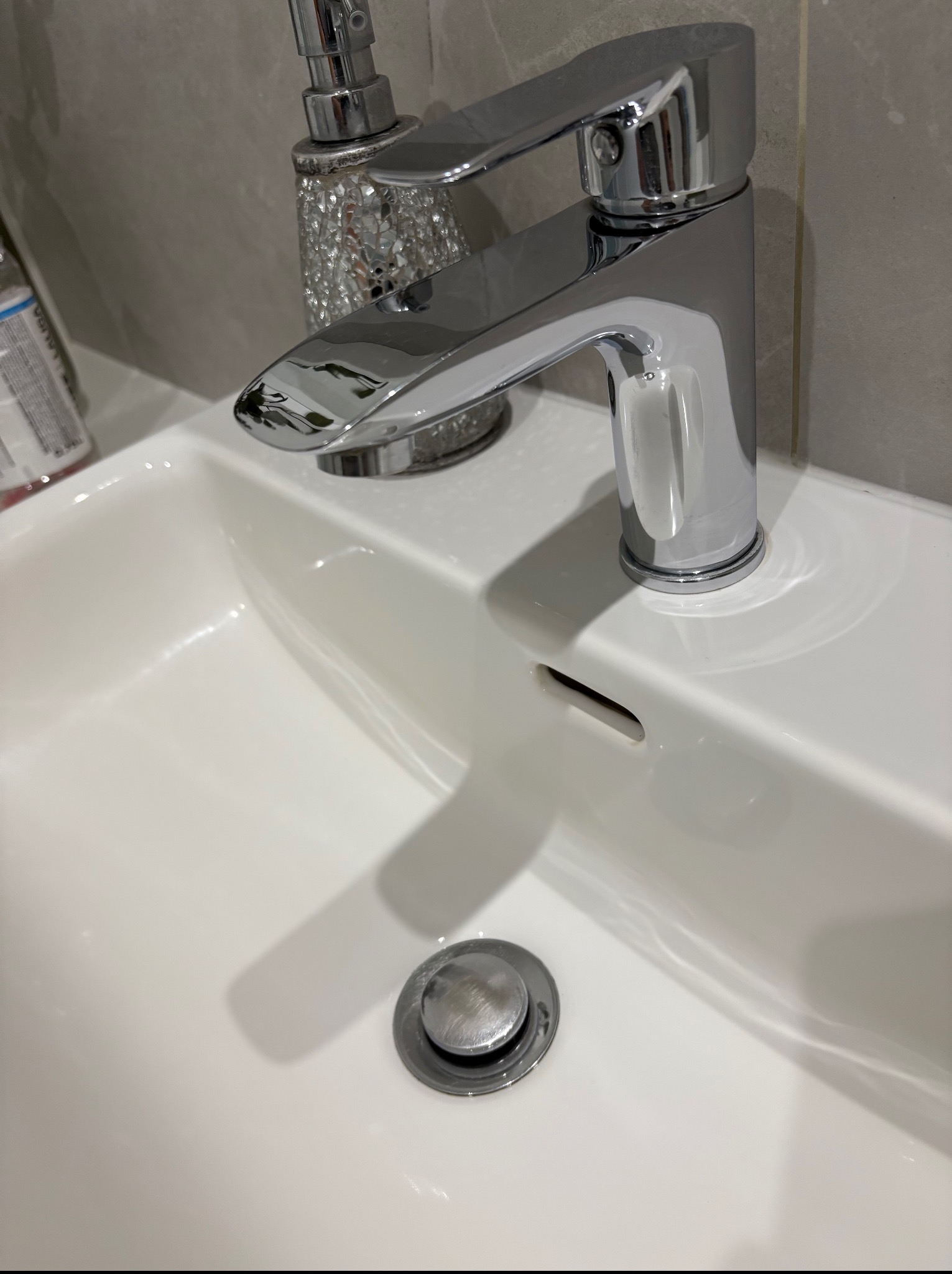 Błyszcząca, chromowana bateria umywalkowa na czystej, białej umywalce z ceramiczną osłoną odpływu. Obok dekoracyjny dozownik mydła.