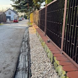 Piotr Babiarz - Ułożona warstwa białego grysu oddzielająca chodnik od ogrodzenia z ceglaną podmurówką. Widoczna czarna agrowłóknina. Tło: ulica z domami i drzewami.