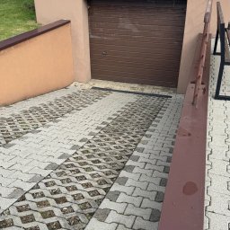 Piotr Babiarz - Podjazd do garażu wyłożony ażurową kostką brukową, częściowo porośniętą trawą. Widoczny fragment budynku z brązową bramą garażową i metalowa barierka.