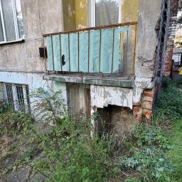 Piotr Babiarz - Zniszczony balkon z odpadającym tynkiem i zardzewiałą balustradą, porośnięty roślinnością. Widoczne uszkodzenia elewacji i zaniedbany teren wokół budynku.