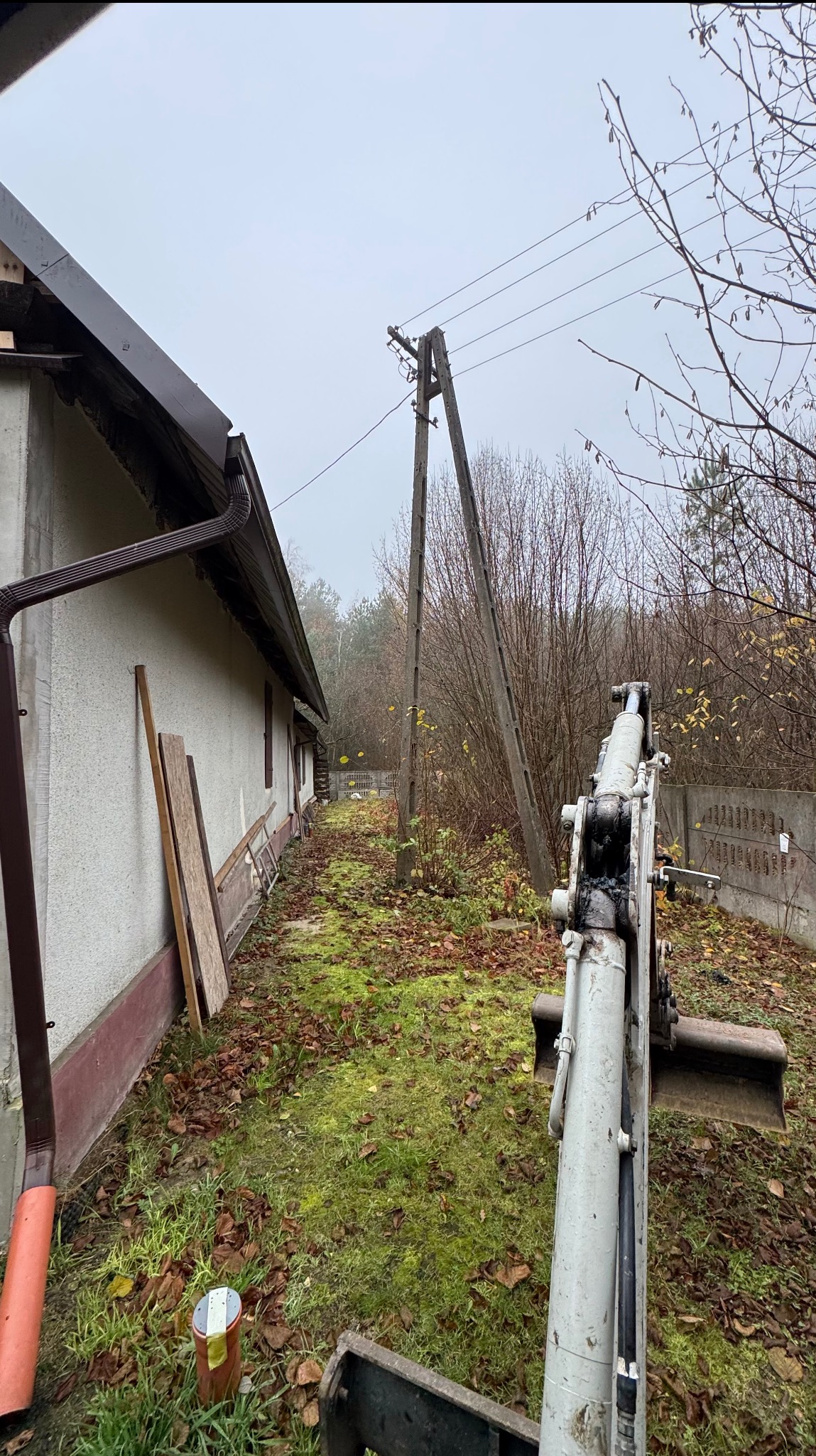 Mini koparka na działce przy domu, przygotowanie terenu pod ogród. Widoczne drzewa i trawa. Prace ziemne w toku, jesienna aura.