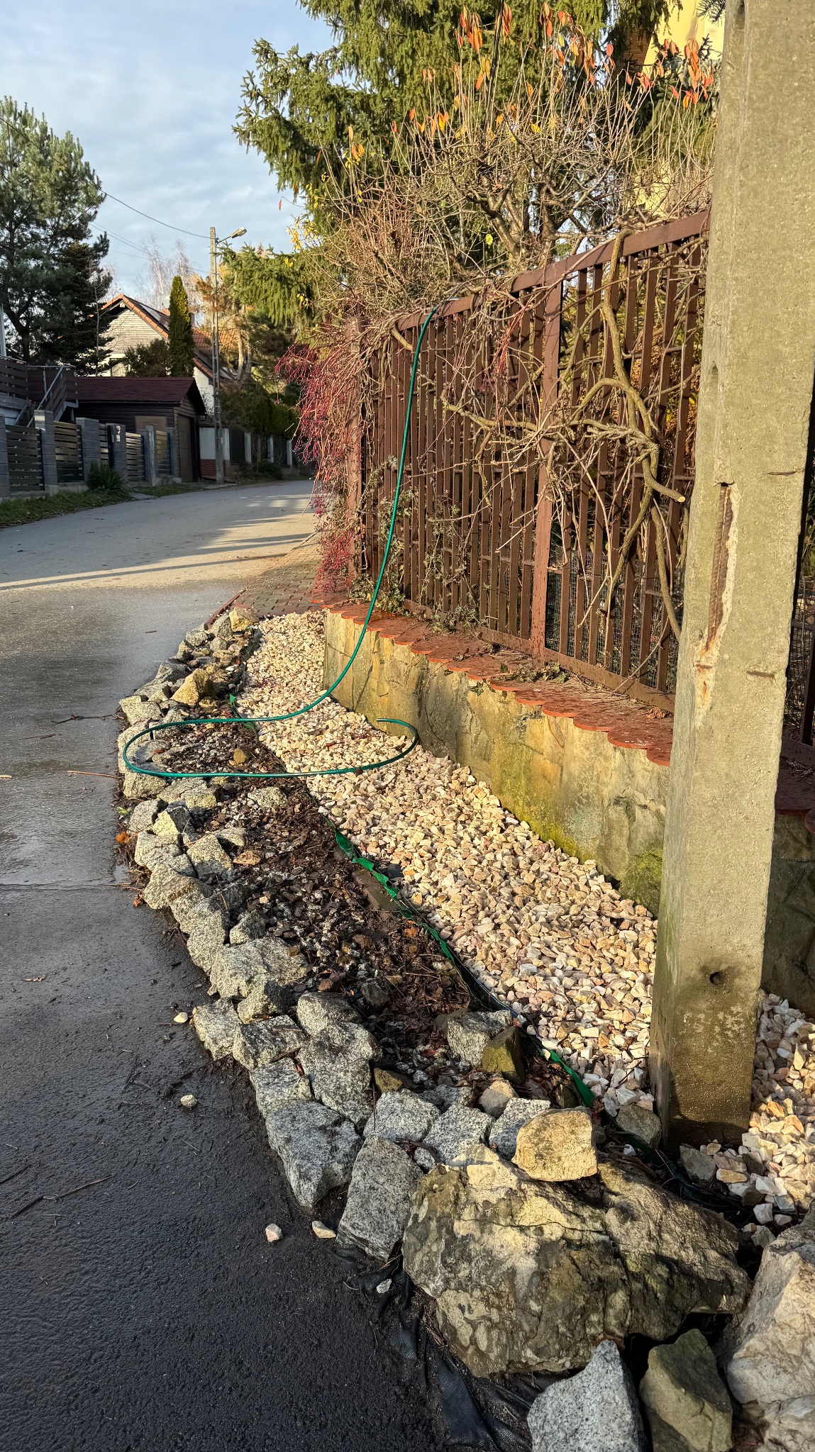 Rabata z kamieniami i korą, oddzielona od chodnika kamieniami. W tle brązowe ogrodzenie z pnączami i zielony wąż ogrodowy. Słup betonowy po prawej stronie.