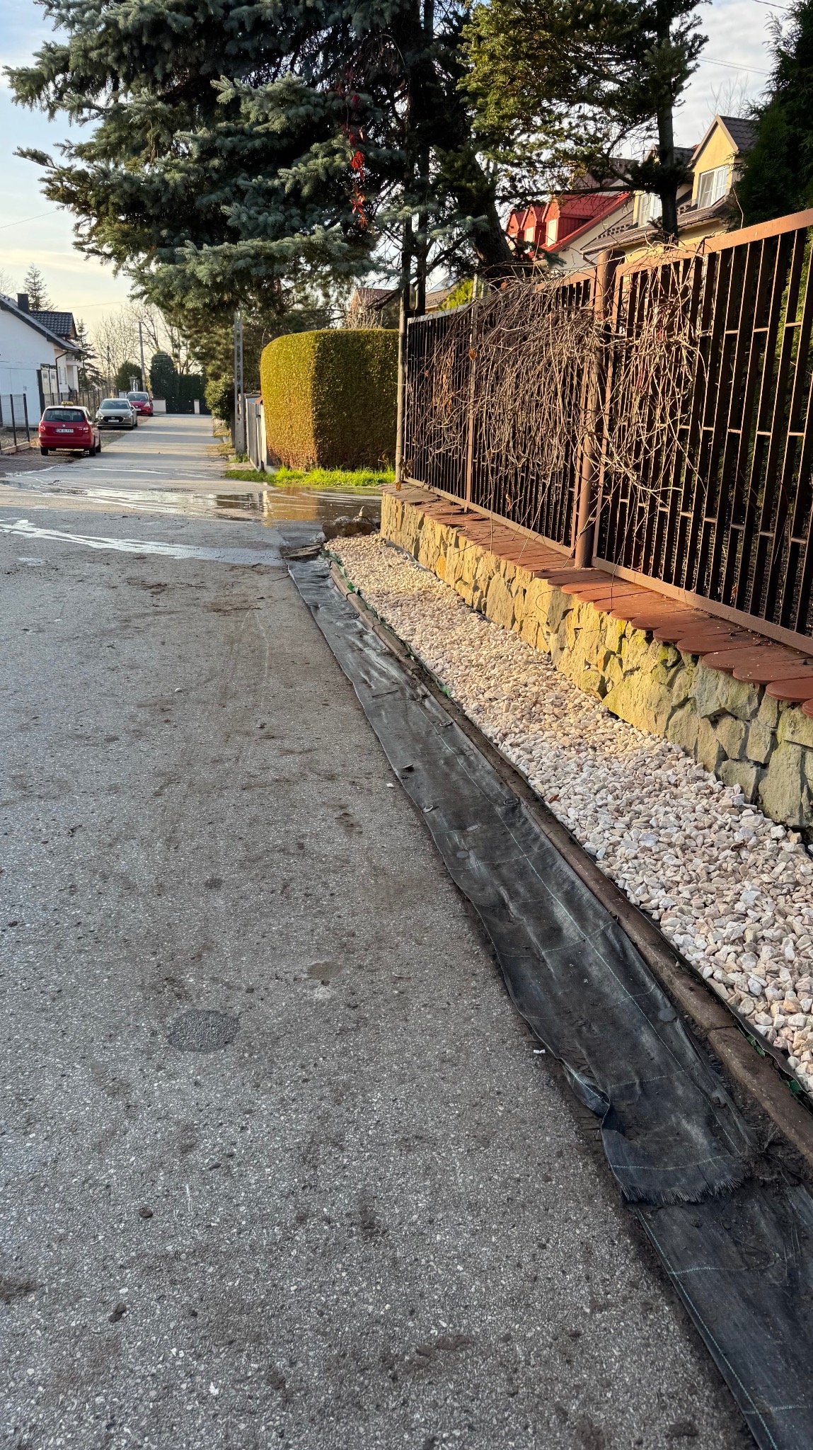 Przygotowanie rabaty z agrowłókniną i białym grysem wzdłuż ogrodzenia na ulicy osiedlowej. W tle domy i drzewa w słoneczny dzień.