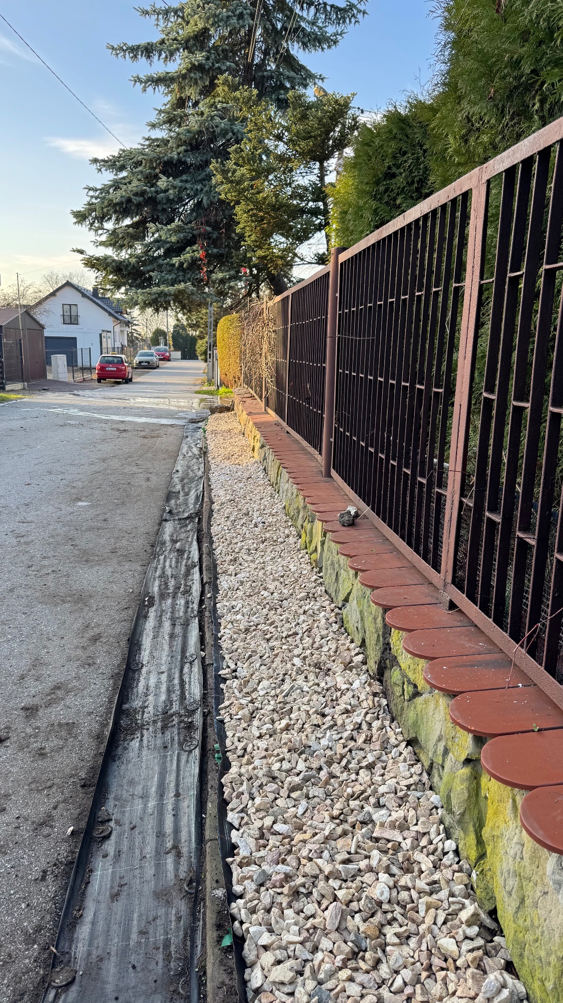Ułożona warstwa białego grysu oddzielająca chodnik od ogrodzenia z ceglaną podmurówką. Widoczna czarna agrowłóknina. Tło: ulica z domami i drzewami.