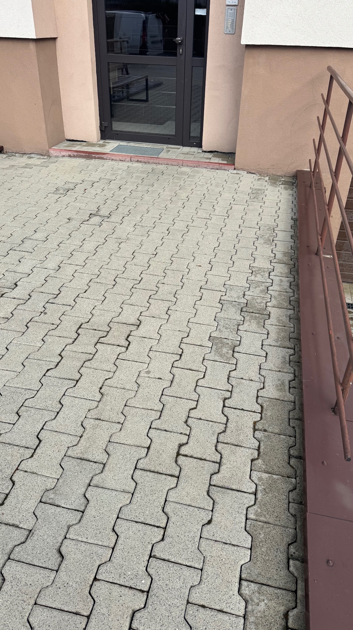 Ułożona szara kostka brukowa przed wejściem do budynku z brązowymi drzwiami i metalową barierką. Widoczny fragment elewacji w odcieniach beżu.