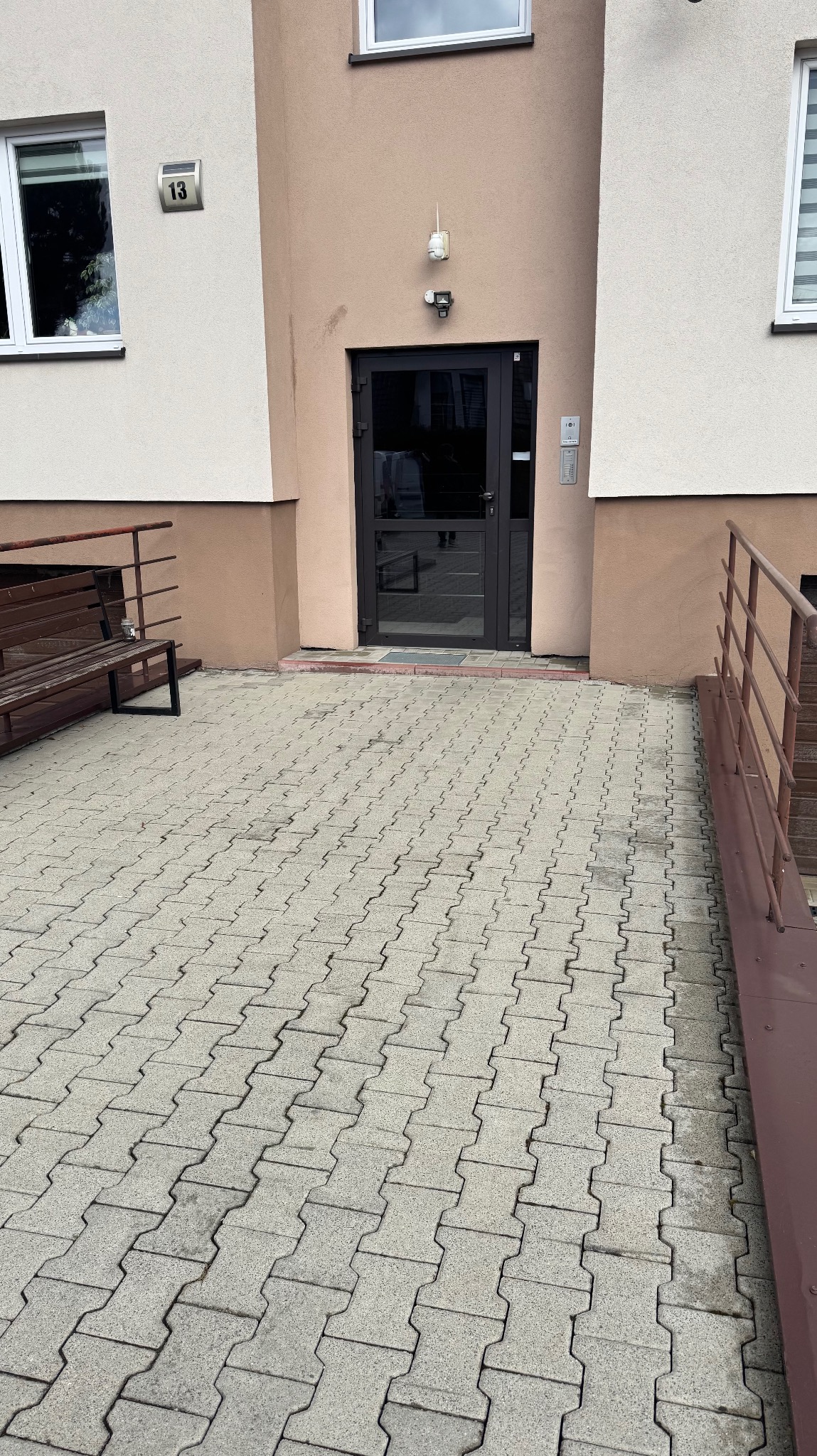 Wejście do budynku z szarej kostki brukowej, ławką i rampą dla niepełnosprawnych. Nad drzwiami kamera monitoringu i numer budynku. Widoczne okna i fragment elewacji.