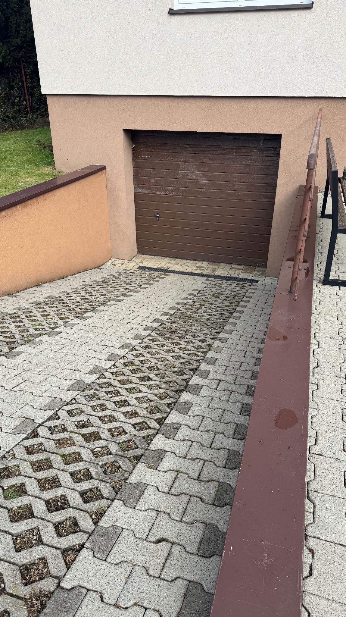 Podjazd do garażu wyłożony ażurową kostką brukową, częściowo porośniętą trawą. Widoczny fragment budynku z brązową bramą garażową i metalowa barierka.