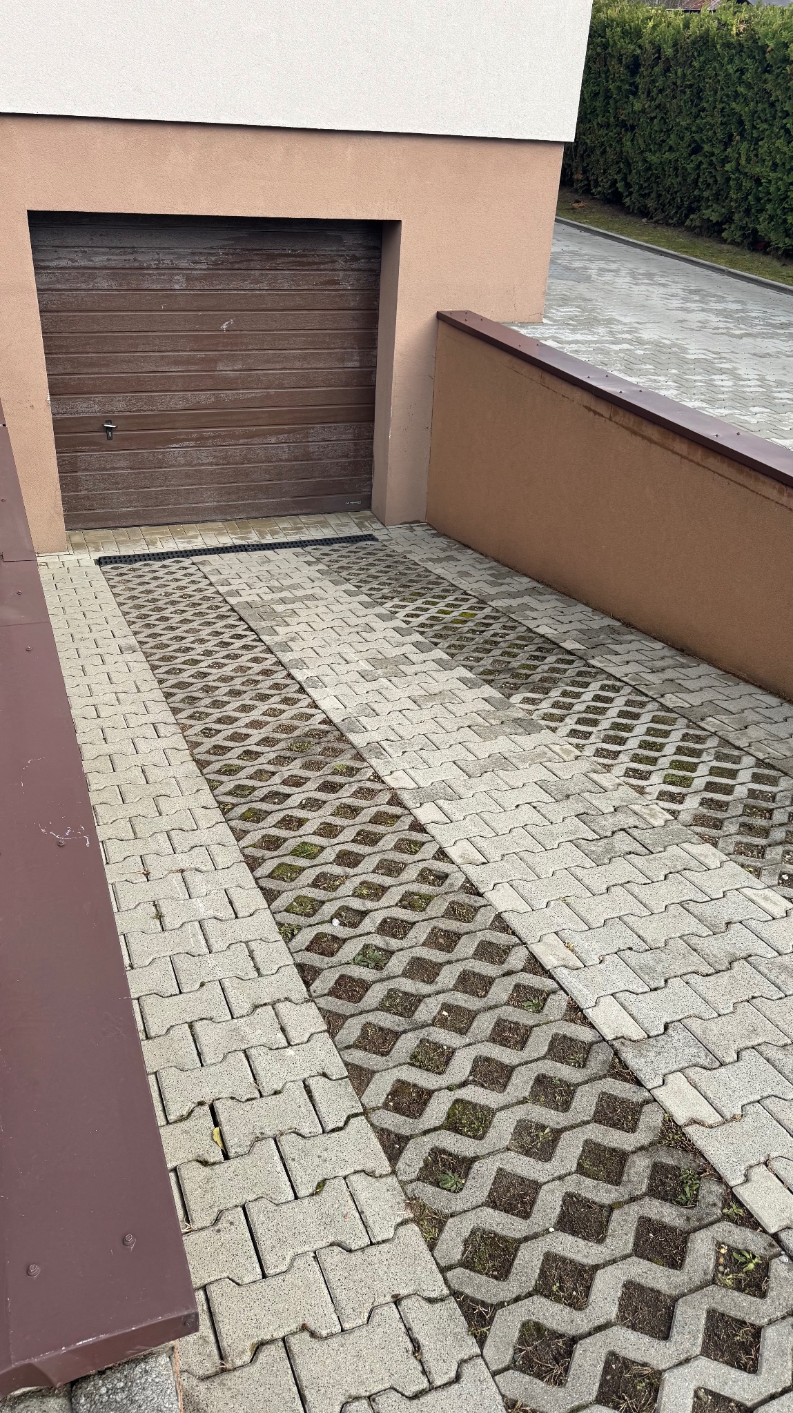Podjazd z ażurowych płyt betonowych prowadzący do garażu z brązową bramą. Widoczne elementy zieleni w przestrzeniach płyt. Ujęcie z góry, lekko z boku.