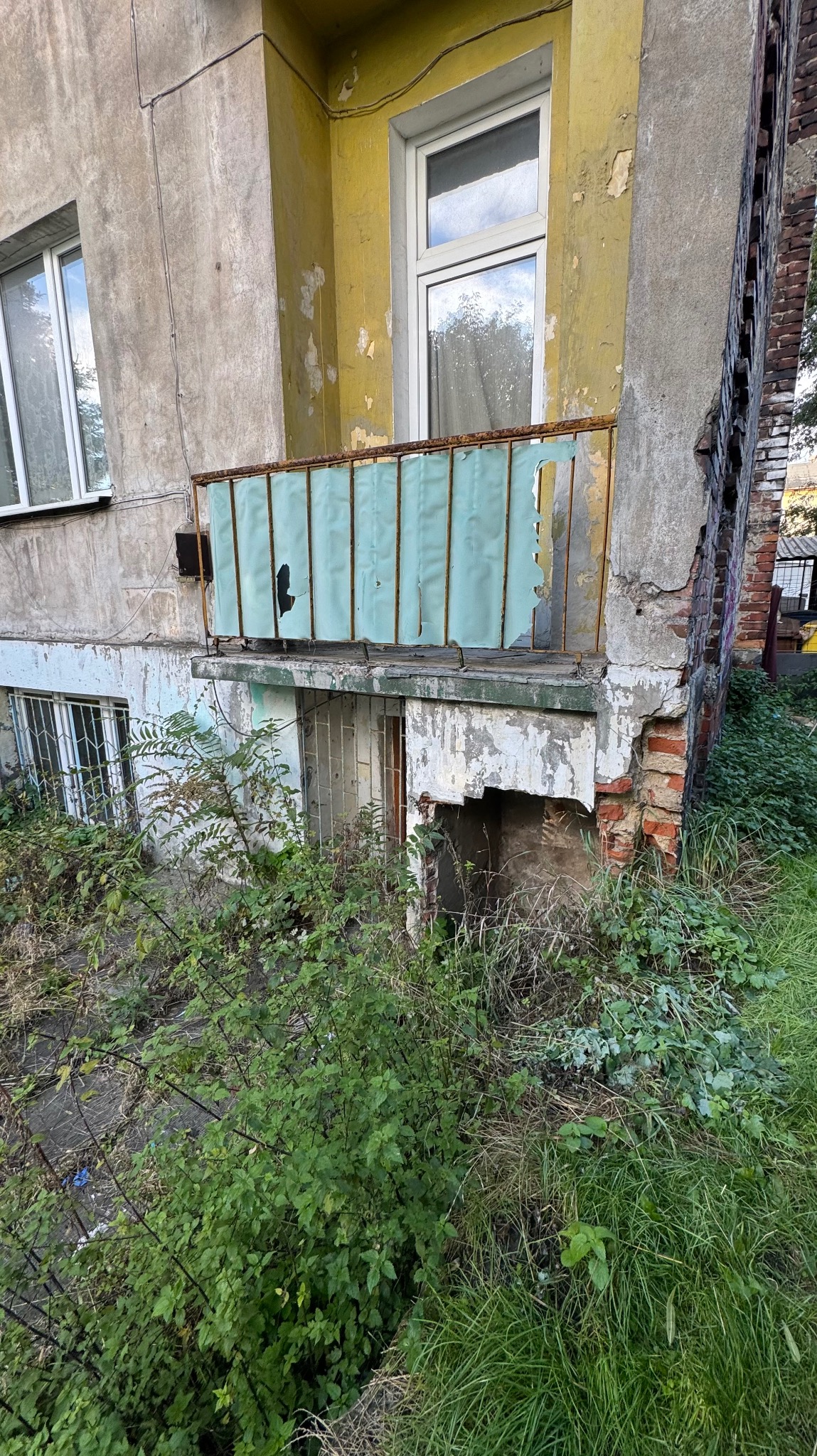 Zniszczony balkon z odpadającym tynkiem i zardzewiałą balustradą, porośnięty roślinnością. Widoczne uszkodzenia elewacji i zaniedbany teren wokół budynku.