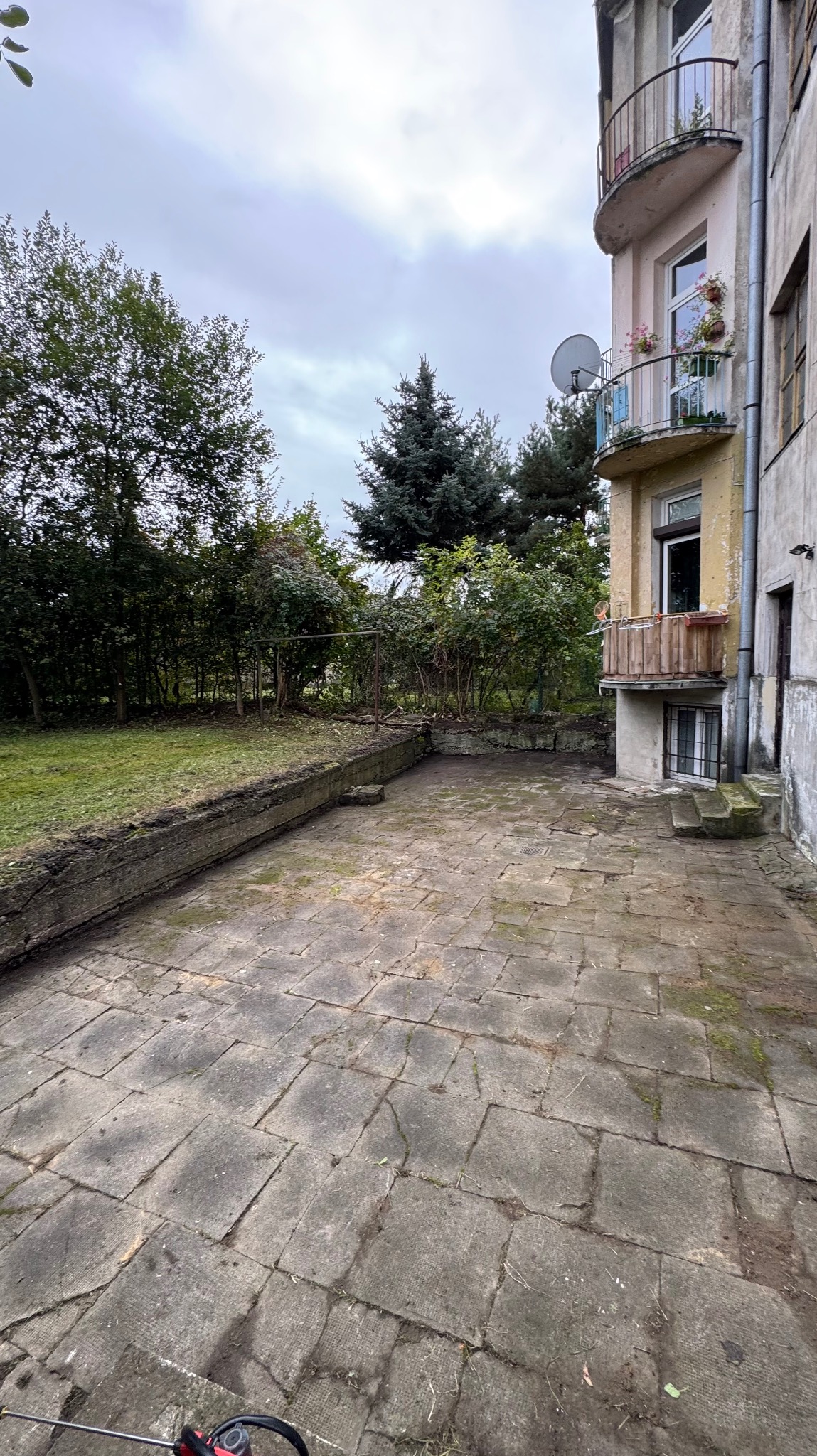 Zaniedbany, kamienny taras przy budynku z balkonami, otoczony zielenią. Widoczne zabrudzenia i porosty na płytach tarasowych, sugerujące potrzebę czyszczenia.