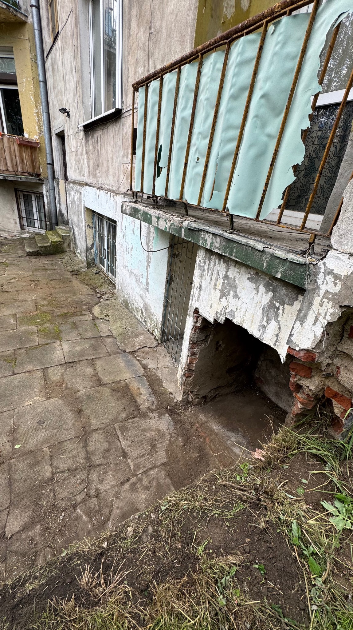 Zniszczony balkon z rdzewiejącą balustradą i podartą osłoną. Widoczne uszkodzenia muru, odpadający tynk i zaniedbany teren wokół budynku.