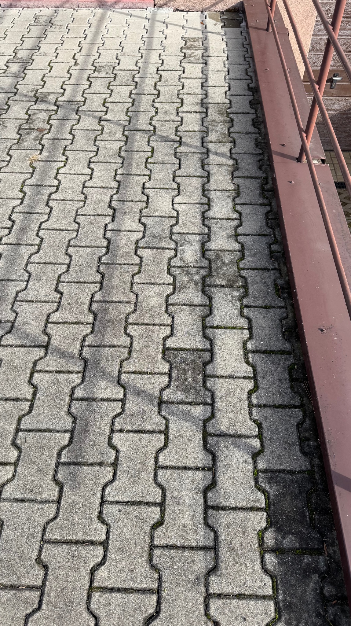 Szara kostka brukowa typu I na zewnątrz, z widocznymi śladami użytkowania i mchem w spoinach, częściowo zacieniona, z fragmentem brązowej barierki po prawej stronie.