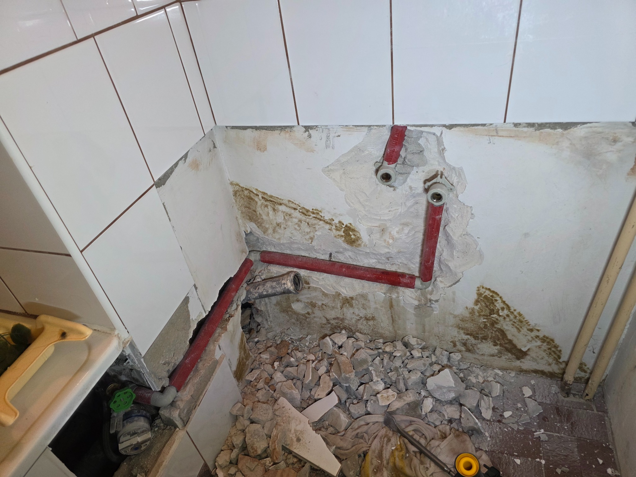 Częściowo wykończona instalacja wodna z czerwonymi rurami w rogu łazienki w trakcie remontu, z widocznymi gruzami i odkrytymi fragmentami ścian.