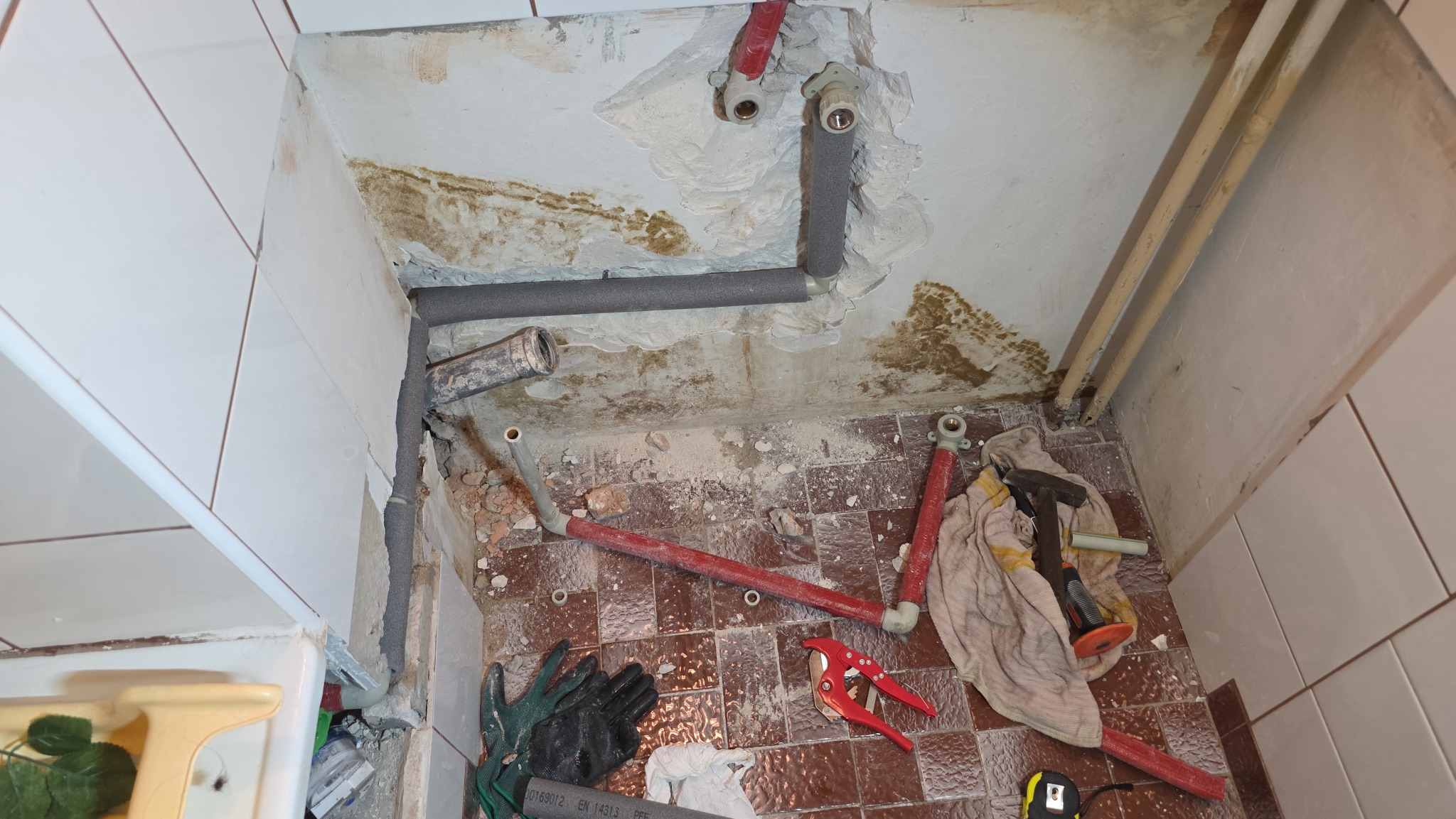 Renowacja łazienki: widok na instalację hydrauliczną w trakcie prac. Rury, narzędzia i fragmenty płytek na podłodze. Prace hydrauliczne w trakcie realizacji.