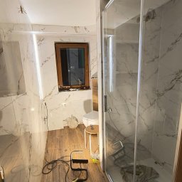 REMBUD PLUS sp. z o.o. - Wykończona łazienka z kabiną prysznicową, marmurowymi płytkami i drewnopodobną podłogą. Widoczne narzędzia i oświetlenie robocze. Na ścianie imitacja owada.