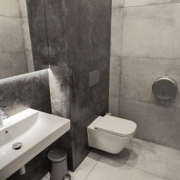 REMBUD PLUS sp. z o.o. - Nowoczesna łazienka z szarymi płytkami, podwieszaną toaletą i umywalką. Minimalistyczny design, oświetlenie LED pod lustrem. Widoczny kosz na śmieci.