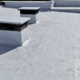 Białe, płaskie pokrycie dachowe z dwoma kominami zabezpieczonymi membraną. Widoczne ślady st&oacute;p na powierzchni dachu i elementy instalacji. Ujęcie z g&oacute;ry.