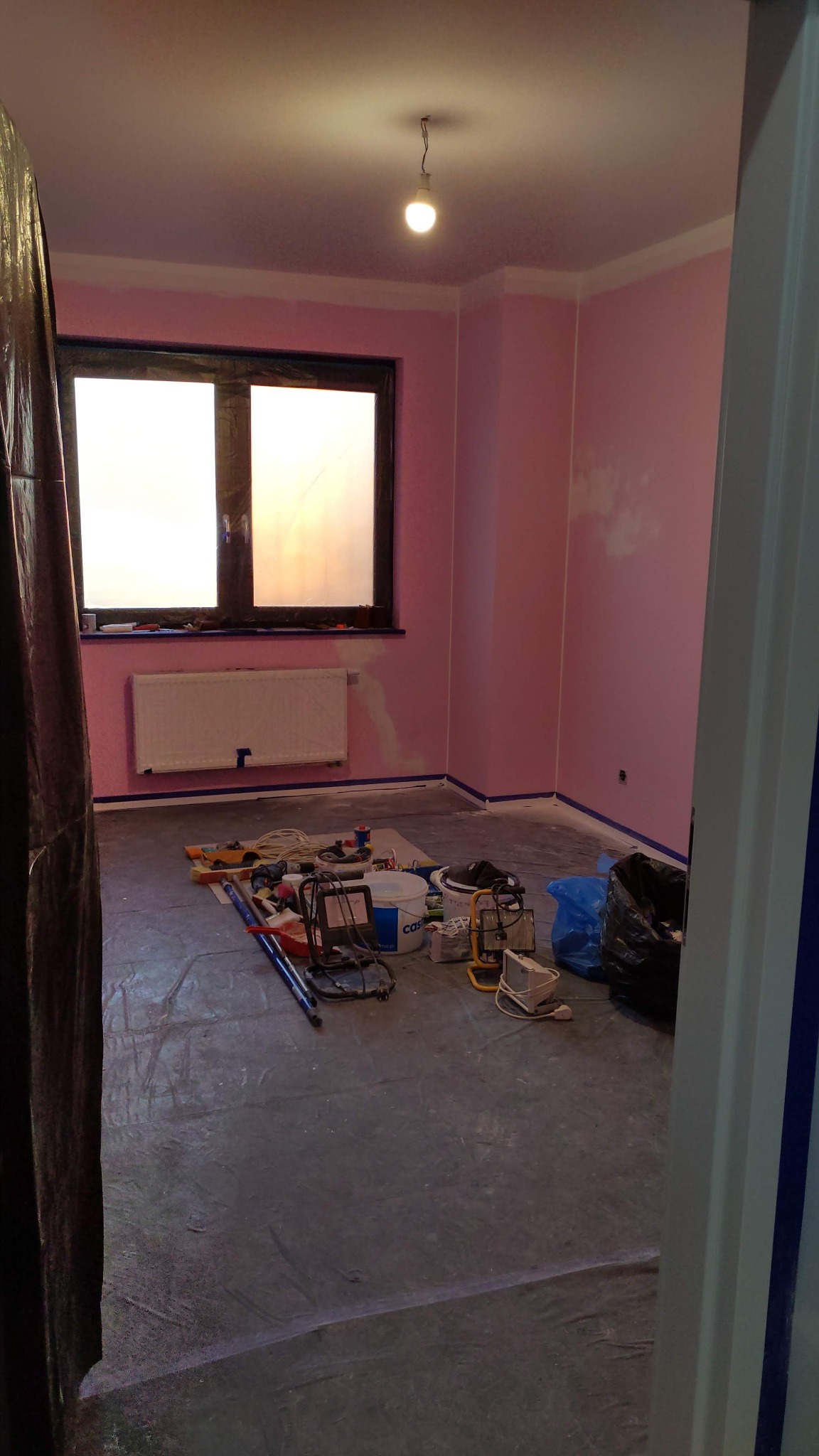 Pokój z różowymi ścianami w trakcie malowania, zabezpieczony folią i taśmą malarską. Widoczne narzędzia malarskie i wiadro z farbą. Przy oknie grzejnik.