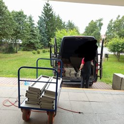 TRANSPORTER ADRIAN RADOMSKI - Wózek z aluminiowymi profilami przed busem z otwartymi drzwiami. Osoba w środku. Teren zielony w tle. Przygotowania do transportu.