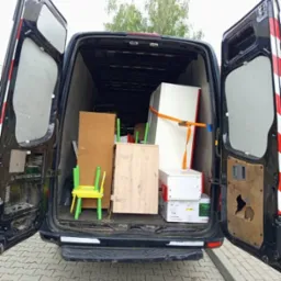 Otwarty bagażnik busa załadowany meblami i kartonami, zabezpieczonymi pasem transportowym. Widoczny fragment chodnika z kostki.