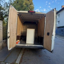 TRANSPORTER ADRIAN RADOMSKI - Wnętrze busa z otwartymi drzwiami, przygotowanego do transportu mebli. Widoczna szafka w środku i zabudowa ścian. Tło: ulica, budynek, drzewo.