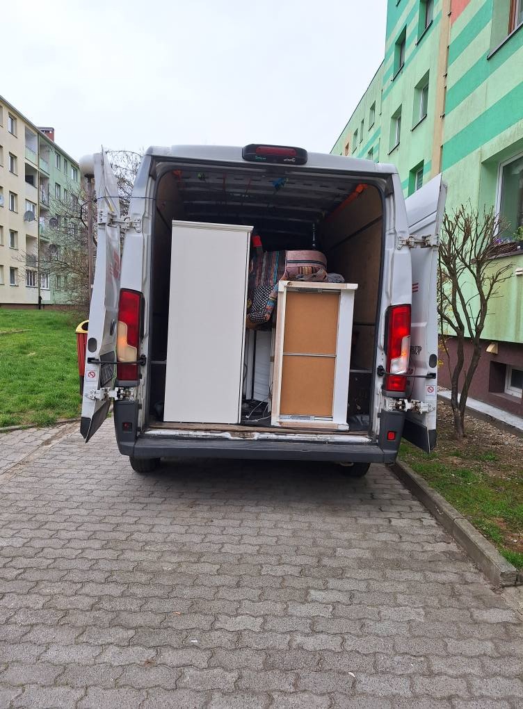 Bus z otwartymi drzwiami załadowany meblami na tle bloku mieszkalnego. Widok z tyłu pojazdu na kostkę brukową.