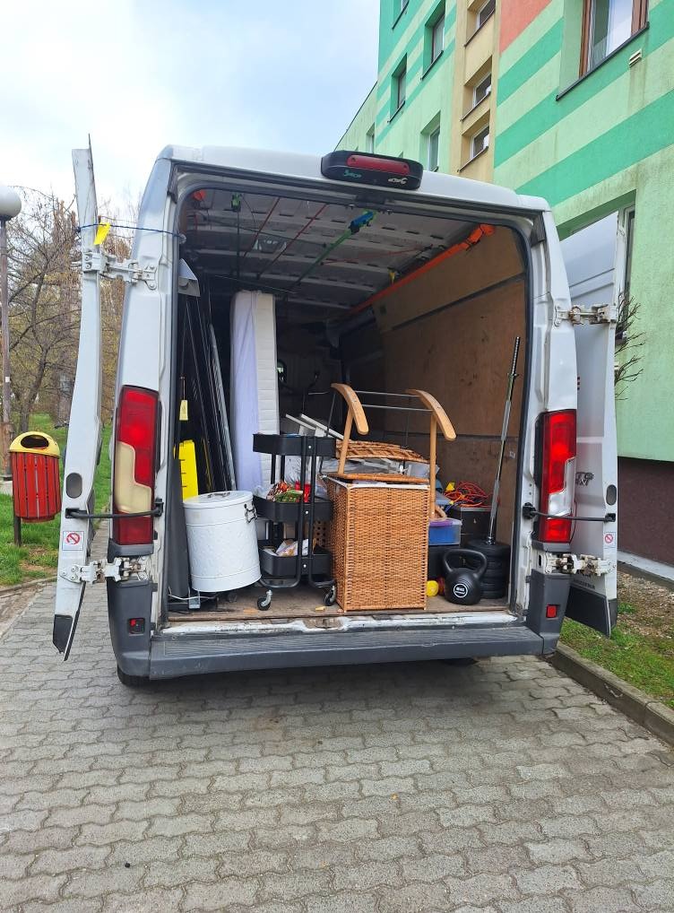 Otwarty bus transportowy z załadunkiem: materac, kosz wiklinowy, kettlebell i wózek na kółkach. Budynek mieszkalny w tle. Widok z tyłu pojazdu.