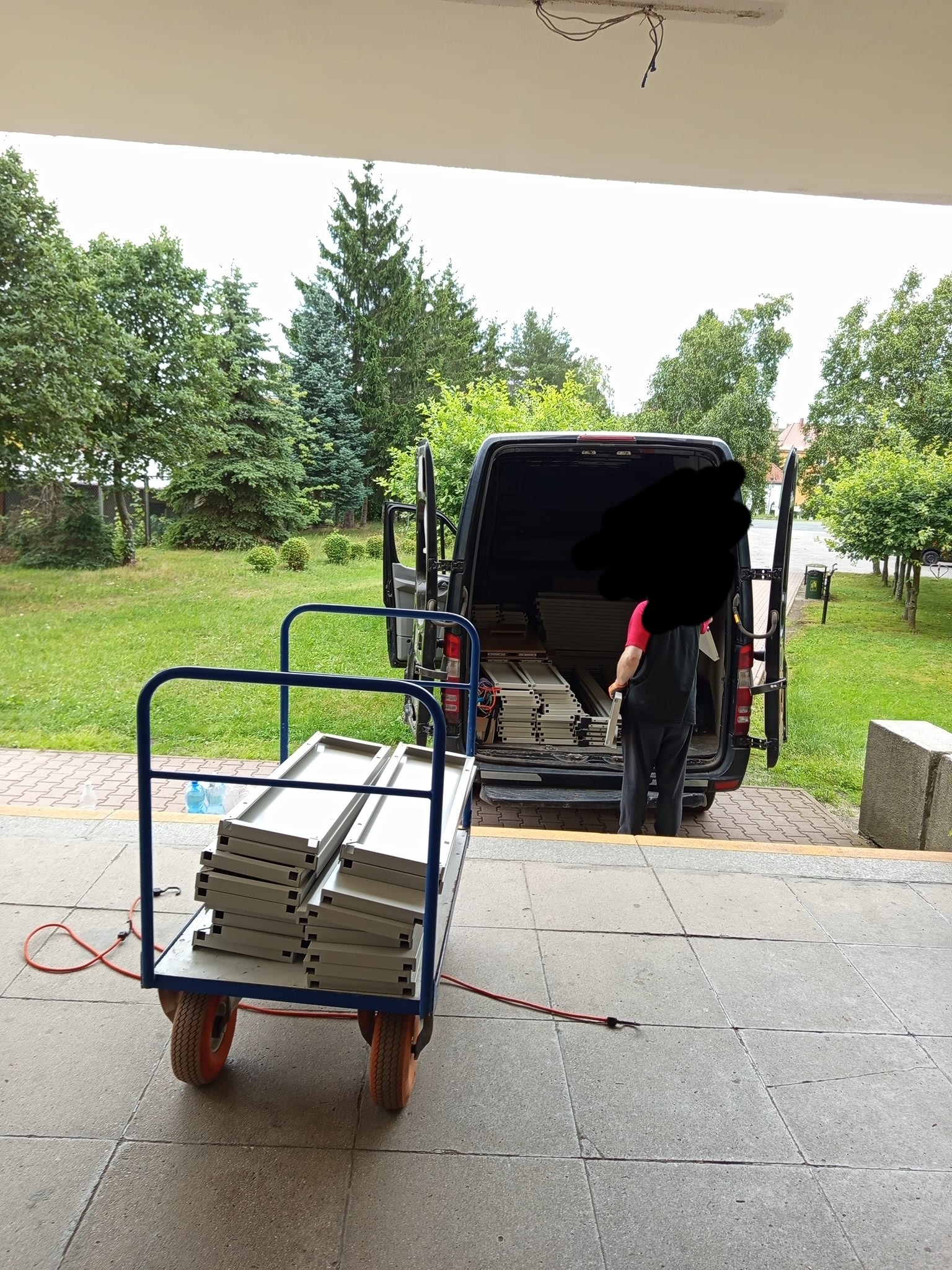 Wózek z aluminiowymi profilami przed busem z otwartymi drzwiami. Osoba w środku. Teren zielony w tle. Przygotowania do transportu.