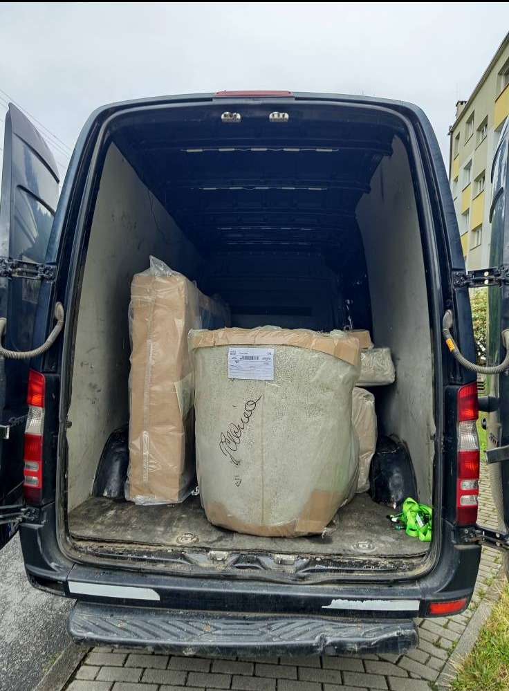 Wnętrze busa załadowane meblami i przedmiotami, przygotowanymi do transportu. Widoczne zabezpieczone paczki i sprzęty. Otwarte drzwi pojazdu.