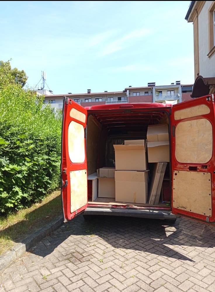 Czerwony bus z otwartymi drzwiami, załadowany kartonami i płytami, gotowy do transportu. Widok z tyłu, na tle zieleni i budynku mieszkalnego.