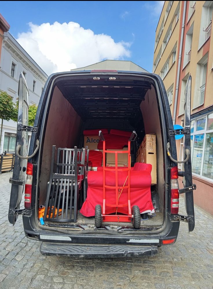 Otwarty tył czarnego busa wypełnionego meblami, krzesłami i czerwonym wózkiem transportowym na tle miejskiej zabudowy. Widoczne kartony i zabezpieczone meble.