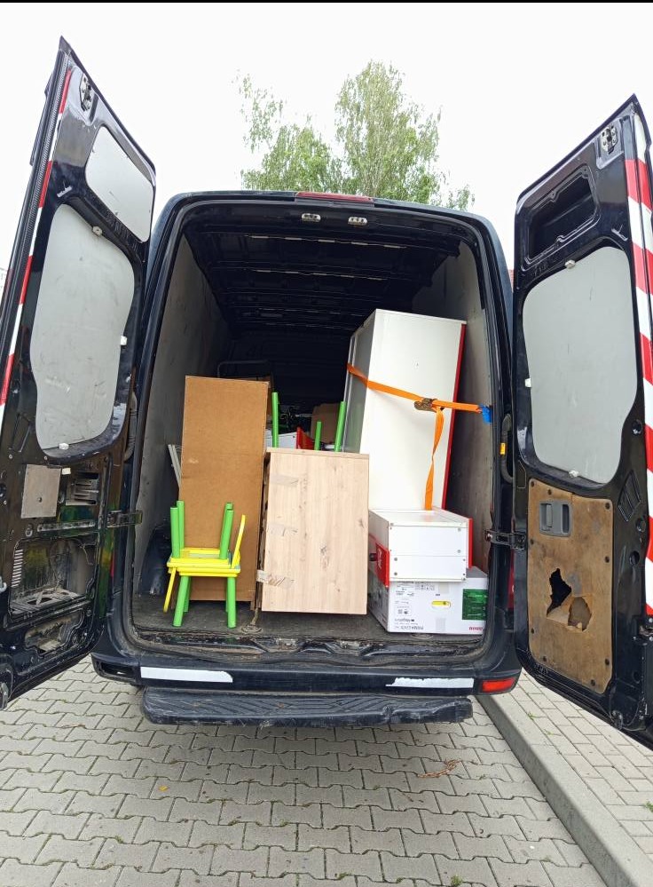 Otwarty bagażnik busa załadowany meblami i kartonami, zabezpieczonymi pasem transportowym. Widoczny fragment chodnika z kostki.