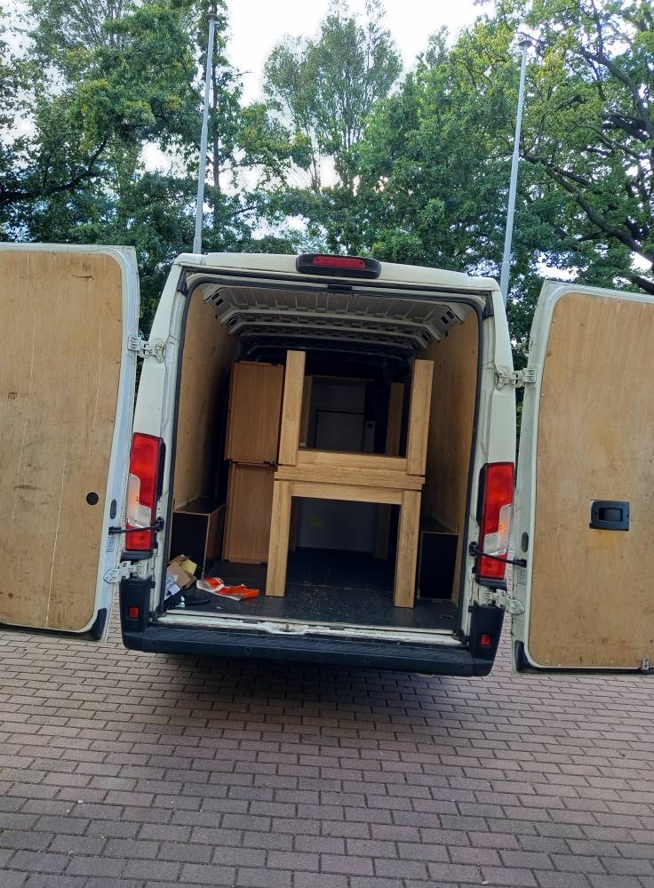 Otwarty tył białego busa załadowanego meblami z jasnego drewna. Widoczne szafki i stół. Na podłodze leżą buty. Tło stanowią drzewa i kostka brukowa.