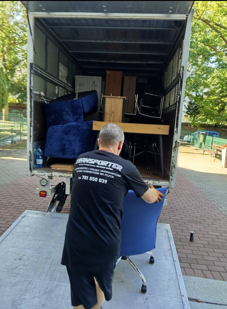 Mężczyzna ładuje niebieskie krzesło do busa transportowego wypełnionego meblami, widok z tyłu, na koszulce logo firmy transportowej.