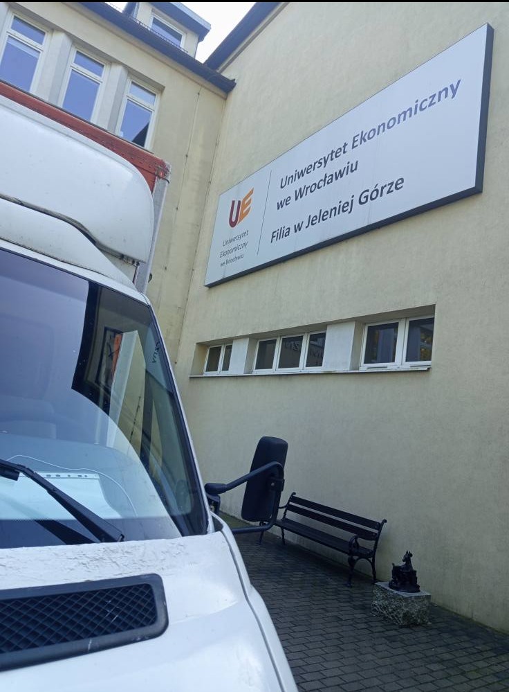 Biały bus przed budynkiem Uniwersytetu Ekonomicznego we Wrocławiu, filia w Jeleniej Górze. Widoczny fragment karoserii i szyby pojazdu, ławka i rzeźba.