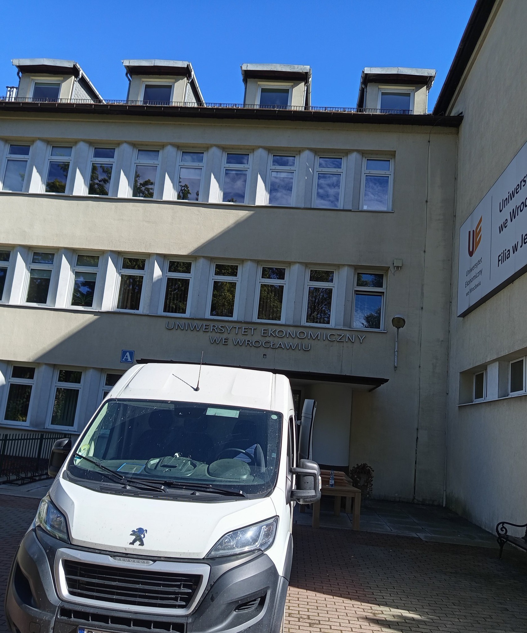 Biały bus Peugeot na tle budynku Uniwersytetu Ekonomicznego we Wrocławiu. Słoneczny dzień, błękitne niebo. Usługa transportowa w przestrzeni miejskiej.