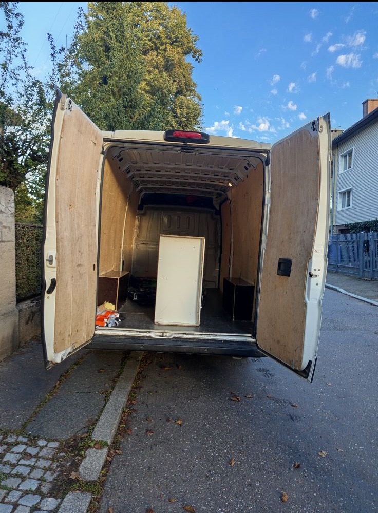 Wnętrze busa z otwartymi drzwiami, przygotowanego do transportu mebli. Widoczna szafka w środku i zabudowa ścian. Tło: ulica, budynek, drzewo.