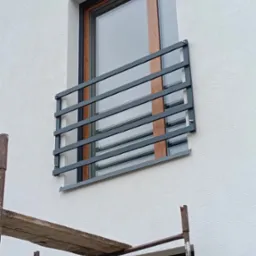 Okna z drewnopodobną ramą i szarymi, poziomymi balustradami zamontowanymi na elewacji budynku. Fragment rusztowania w kadrze.