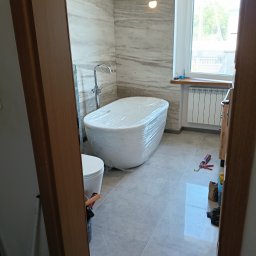 Przedsiębiorstwo Usługowo-Montażowe GoodJob Paweł Cieślik - Nowa, owinięta folią wanna w łazience z szarą marmurową ścianą i oknem. Widok zza drewnianej ościeżnicy. Ślady prac wykończeniowych.