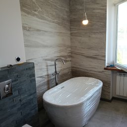 Przedsiębiorstwo Usługowo-Montażowe GoodJob Paweł Cieślik - Nowoczesna łazienka w trakcie wykończenia: wanna owinięta folią ochronną, szara ściana z kamienia, bateria stojąca i okno z grzejnikiem. Widoczna surowa żarówka.