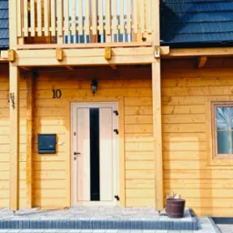Drewniany dom z balkonem, jasnymi drzwiami z czarnym paskiem i numerem 10. Widoczne schody z kostki brukowej i metalowa skrzynka na listy.