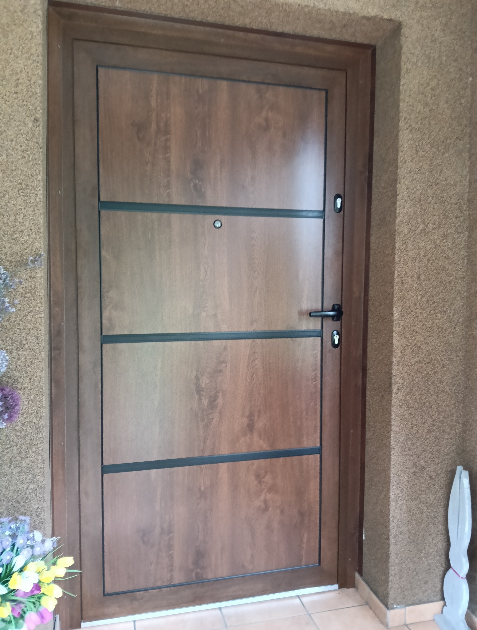 Brązowe drzwi wejściowe z poziomymi, czarnymi wstawkami, zamontowane w budynku o beżowej elewacji. Po lewej stronie widoczny bukiet kwiatów.